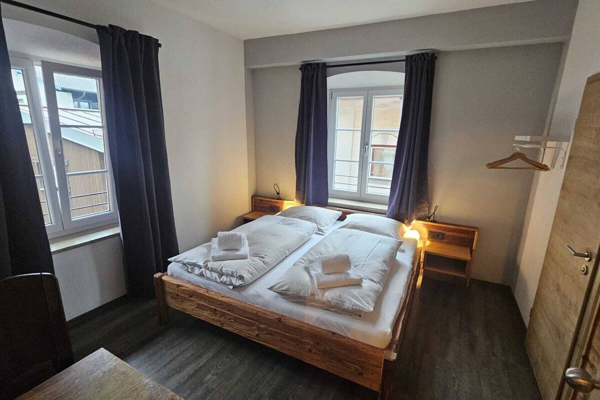 A bedroom with a wooden bed, two bedside tables, a wardrobe, and two windows with curtains.