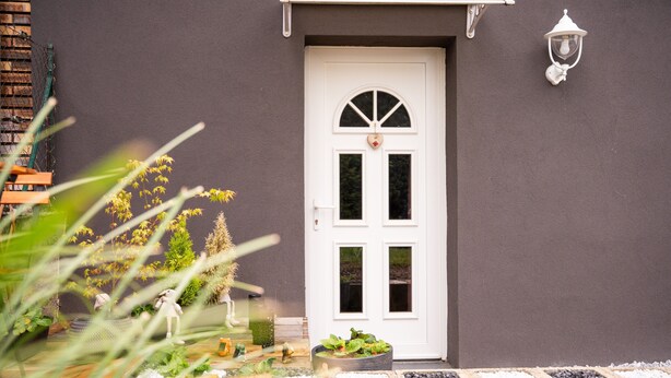 Une porte blanche ornée d’un motif en forme de cœur, un luminaire blanc fixé au mur et un petit espace jardin avec des plantes et une statue d’oiseau décorative.
