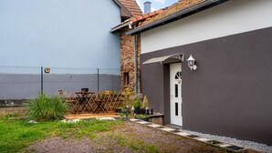 Une maison à l’extérieur grise, avec une porte blanche et un petit jardin clos, comprenant une table en bois et des chaises.