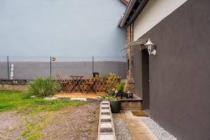 Ein umzäunter Garten mit Terrasse, Gartenmöbeln, Topfpflanzen und einer Wandleuchte.