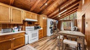 A cabin interior with wooden walls and ceiling, a kitchen with white appliances, a dining area with a table and chairs, and a large window providing natural light. - Mama Bear's Cozy Cabin (Sugarloaf)