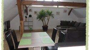 Een gezellige kamer met een houten plafond, een eettafel met stoelen, een potplant, een bank en een televisie op een standaard.