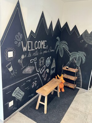 Un mur noir servant de tableau, sur lequel est écrit un message de bienvenue et sont dessinés divers motifs, un banc en bois et un éléphant en jouet.