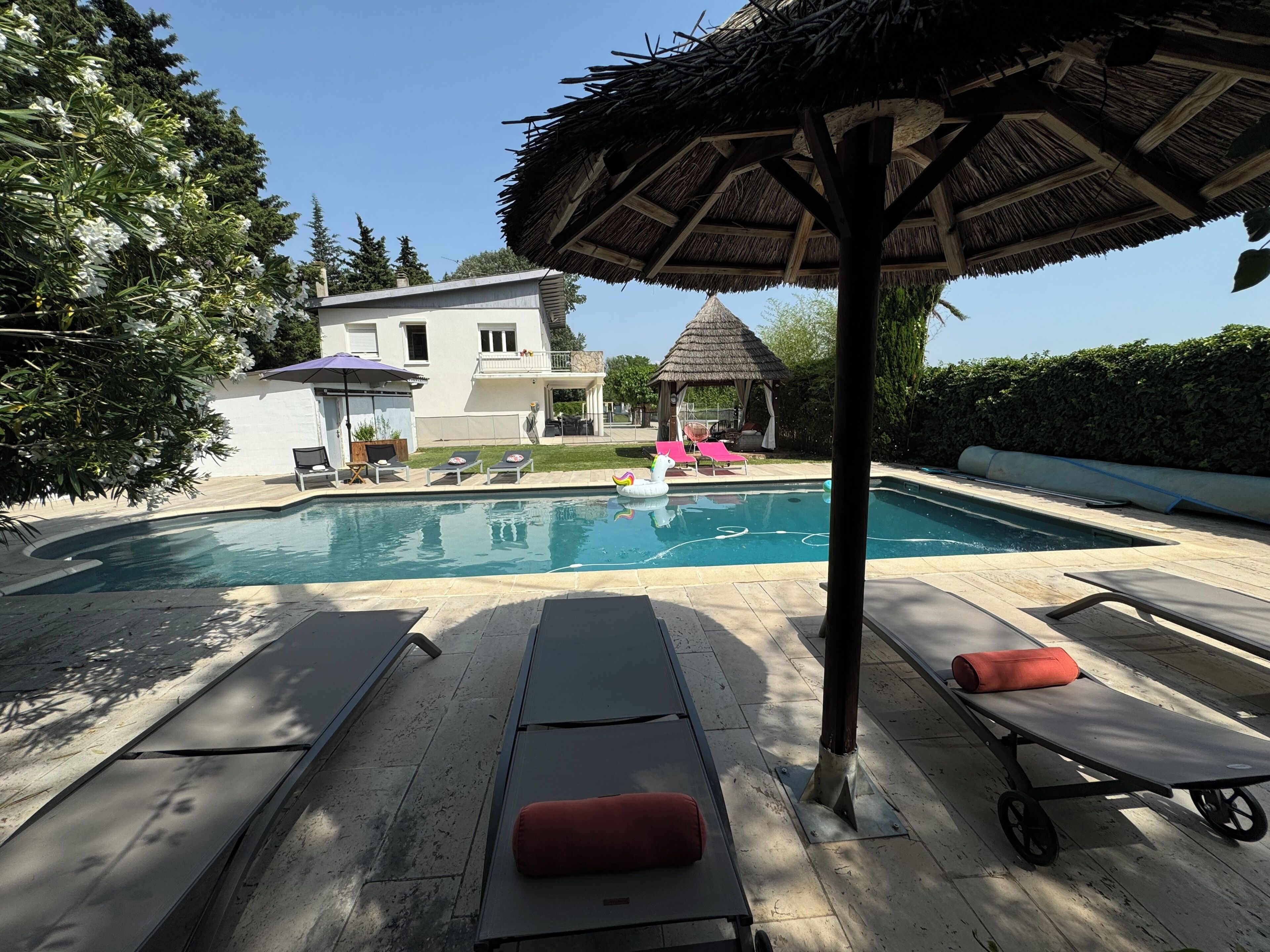 Une piscine avec des chaises longues, un parasol en chaume et une maison en arrière-plan.