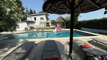 Une piscine avec des chaises longues, un parasol en chaume et une maison en arrière-plan.