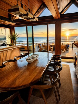 A wooden dining table with chairs, a large window overlooking the ocean, and a view of the sunset.