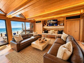 Amplia sala de estar con un televisor grande, techo de madera y vista a la playa.