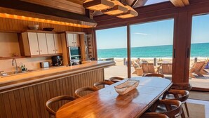 Una zona de bar de madera con vista al océano, una larga mesa de comedor con sillas y un área de estar frente a la playa.