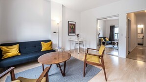 Een moderne woonkamer met een bank, salontafel en stoelen, een eethoek met een tafel en stoelen, en een keuken die zichtbaar is door een open deur.