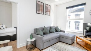 Ein modernes Wohnzimmer mit einem grauen Ecksofa, einem kleinen Beistelltisch, einem Fernseher und einem Fenster mit Blick auf Gebäude.