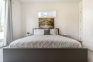 A bedroom with a bed, a window, a painting, and a small table with a plant.