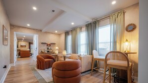 A modern living room with a sofa, ottoman, and wooden chair, a dining area with a table and chairs, and a kitchen in the background. - Renovated Spacious Home Near Downtown, Sleeps 10 (Richmond)