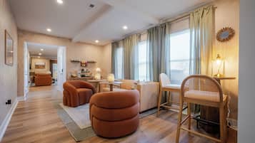 Un salon moderne avec un canapé, un pouf et une chaise en bois, un coin repas avec une table et des chaises, et une cuisine en arrière-plan.