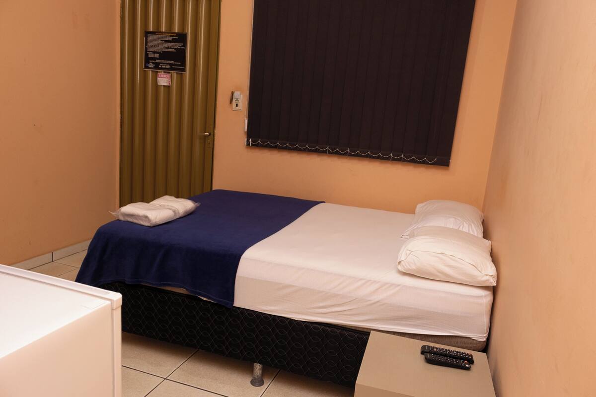 A single bed with a blue and white bedspread, a white pillow, and a remote control on a small table beside the bed.