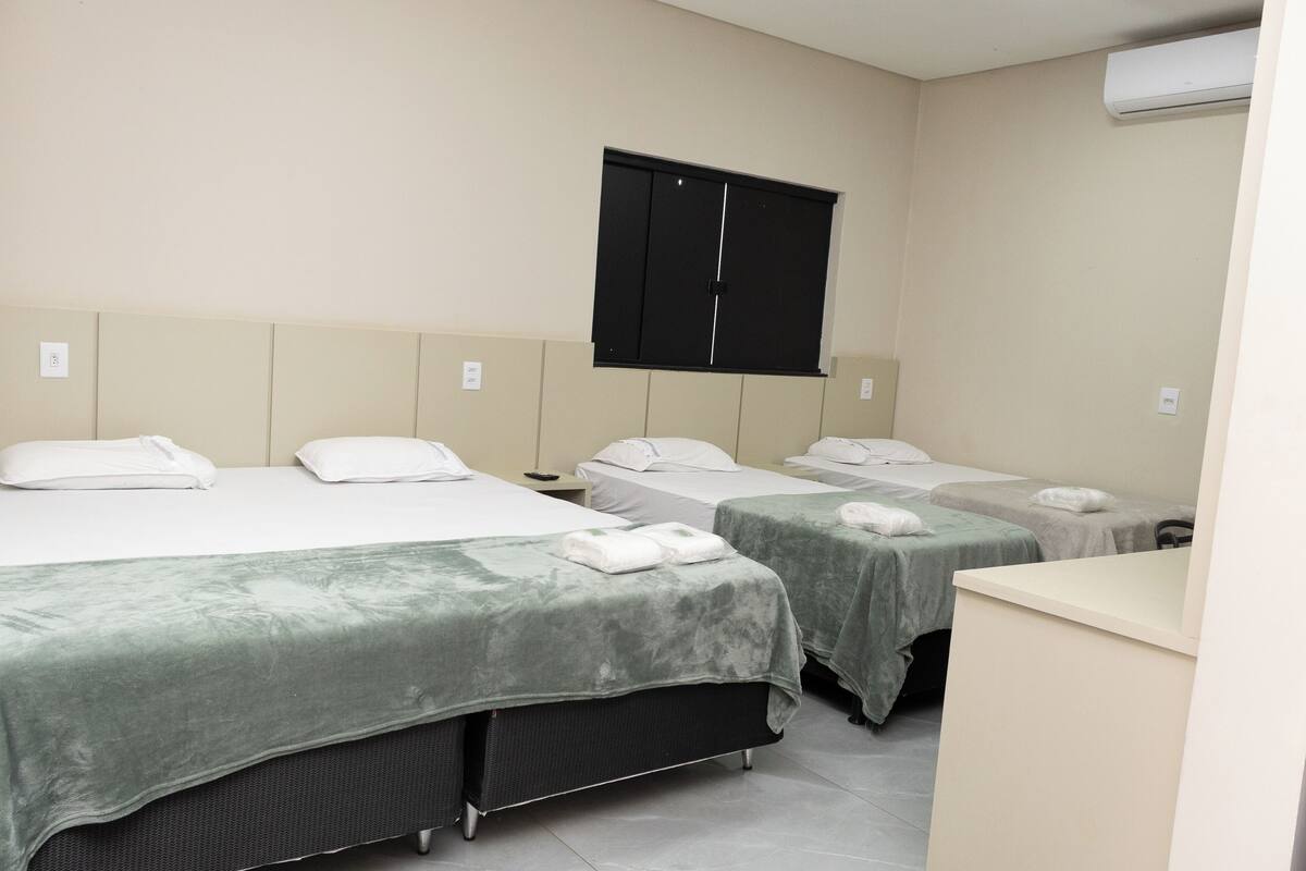 A hotel room with three beds, a black wardrobe, a white air conditioning unit, and a small desk.