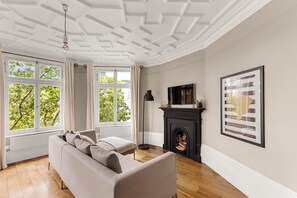 A room with a sofa, a fireplace, and large windows with a view of trees. - Elegant Soho 2-bed, bright Victorian apartment with lift, walk to Covent Garden (London)