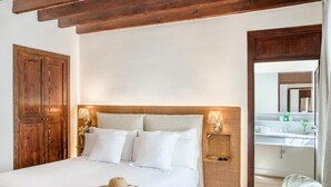 Een slaapkamer met een houten hoofdbord, wit beddengoed, een bedlampje en een strohoed op het bed.