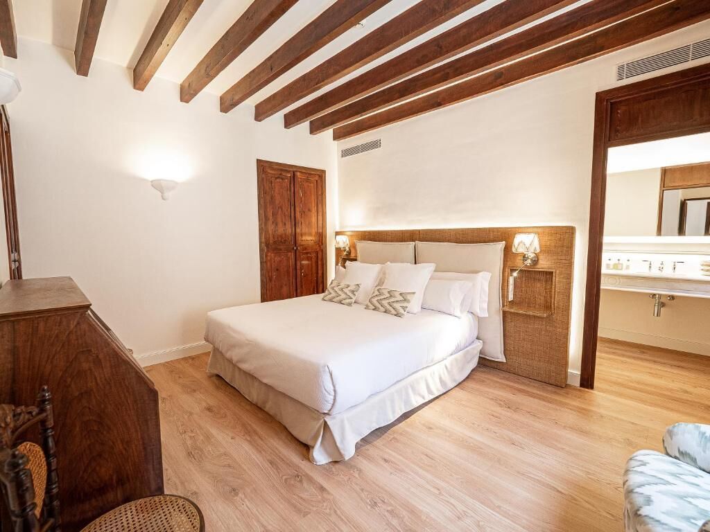 Un dormitorio con techo de madera, una cama con ropa blanca, un aparador de madera y un baño con lavamanos y espejo.
