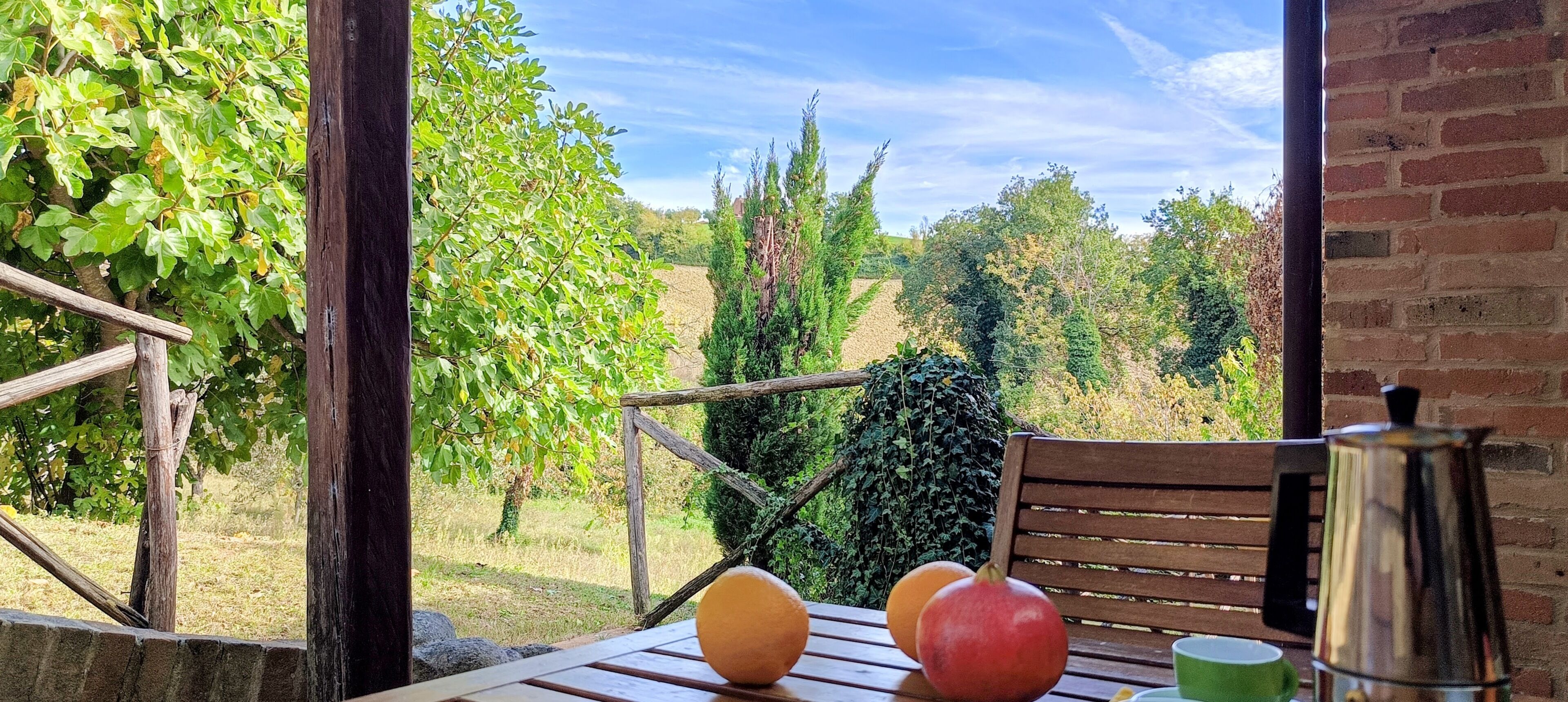 Une table en bois avec des fruits et une cafetière, un mur de briques et une vue sur un jardin avec des arbres et une clôture.