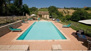 Ein rechteckiger Swimmingpool mit einer gefliesten Liegefläche, umgeben von Liegestühlen und Sonnenschirmen, vor einer Kulisse aus sanften Hügeln und Bäumen.