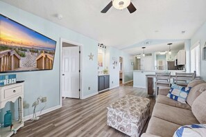 A spacious living room with a large painting, a ceiling fan, and a kitchen in the background. - Waterway Paradise Escape Gated Coastal Retreat (North Myrtle Beach)