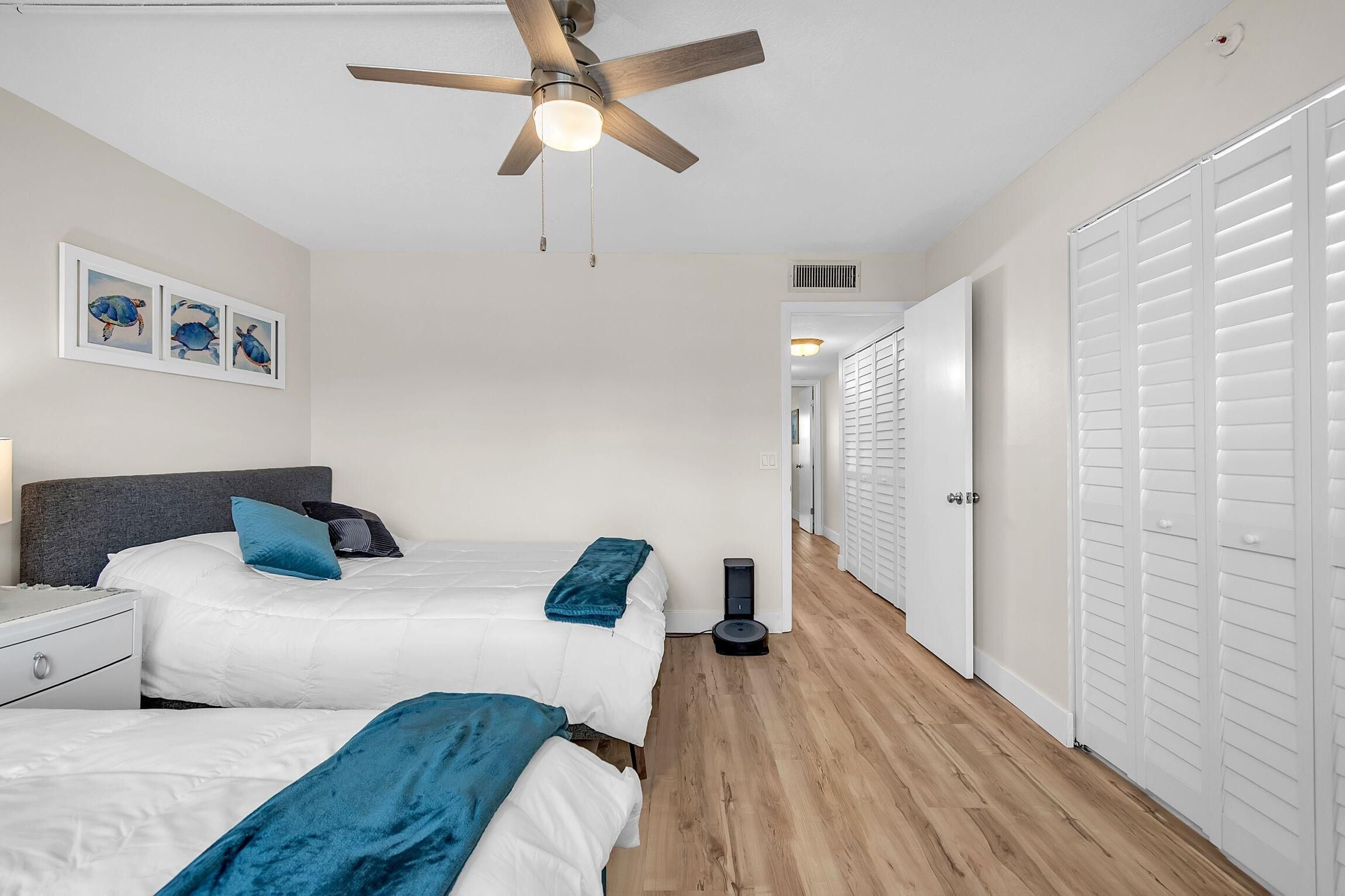 Une chambre avec deux lits, un ventilateur de plafond, un aspirateur et un couloir menant à un placard.
