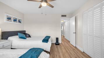 Une chambre avec deux lits, un ventilateur de plafond, un aspirateur et un couloir menant à un placard.