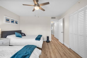 Une chambre avec deux lits, un ventilateur de plafond, un aspirateur et un couloir menant à un placard.