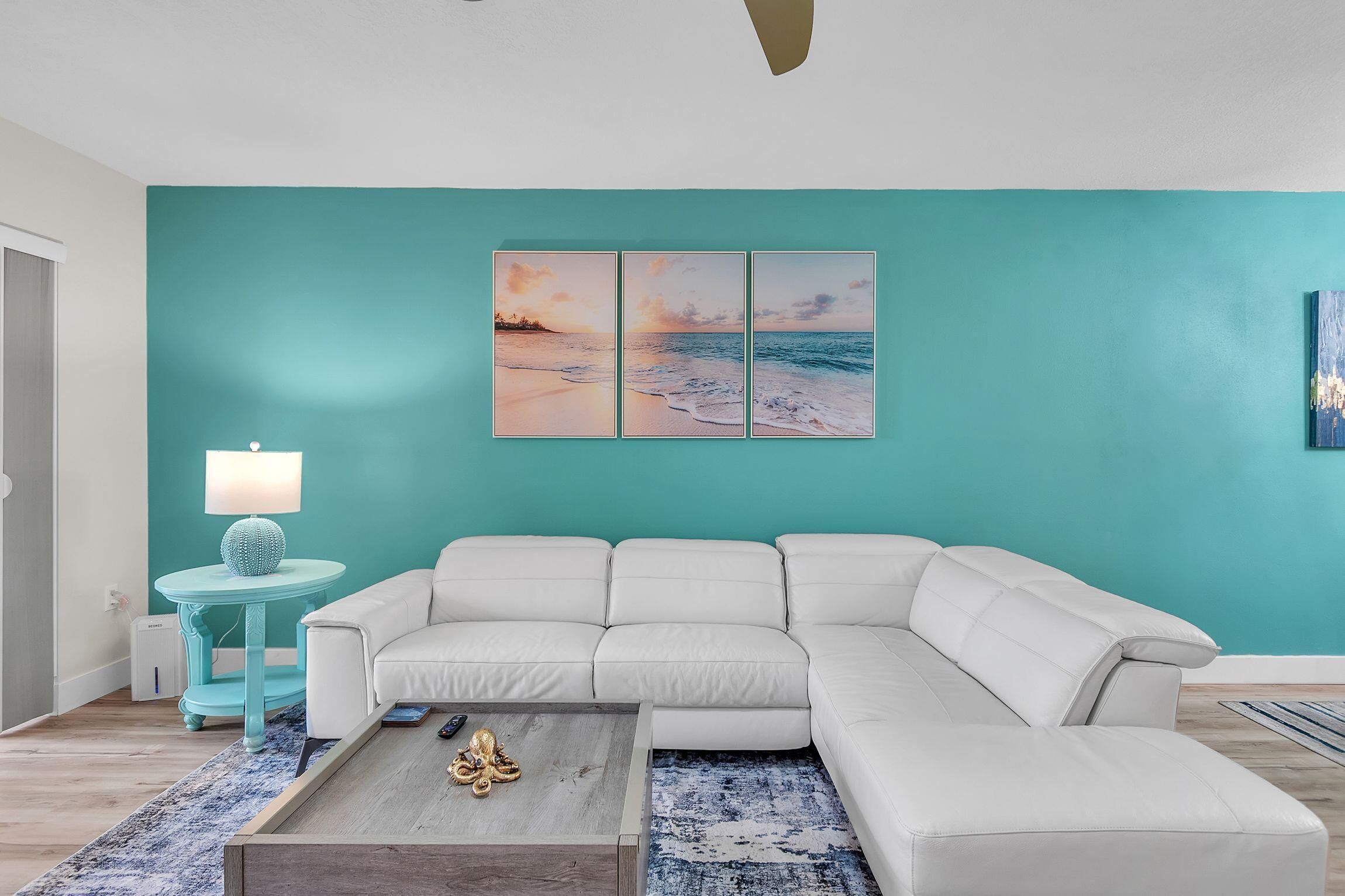 A living room with a white sectional sofa, a teal wall, and a framed beach scene.
