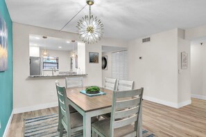 A dining area with a wooden table, white chairs, a hanging light fixture, and a kitchen visible through an open doorway. - Golden Shores #210 (Indian Shores)