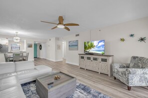 A modern living room with a white sofa, a coffee table, a TV on a cabinet, and a ceiling fan. - Golden Shores #210 (Indian Shores)