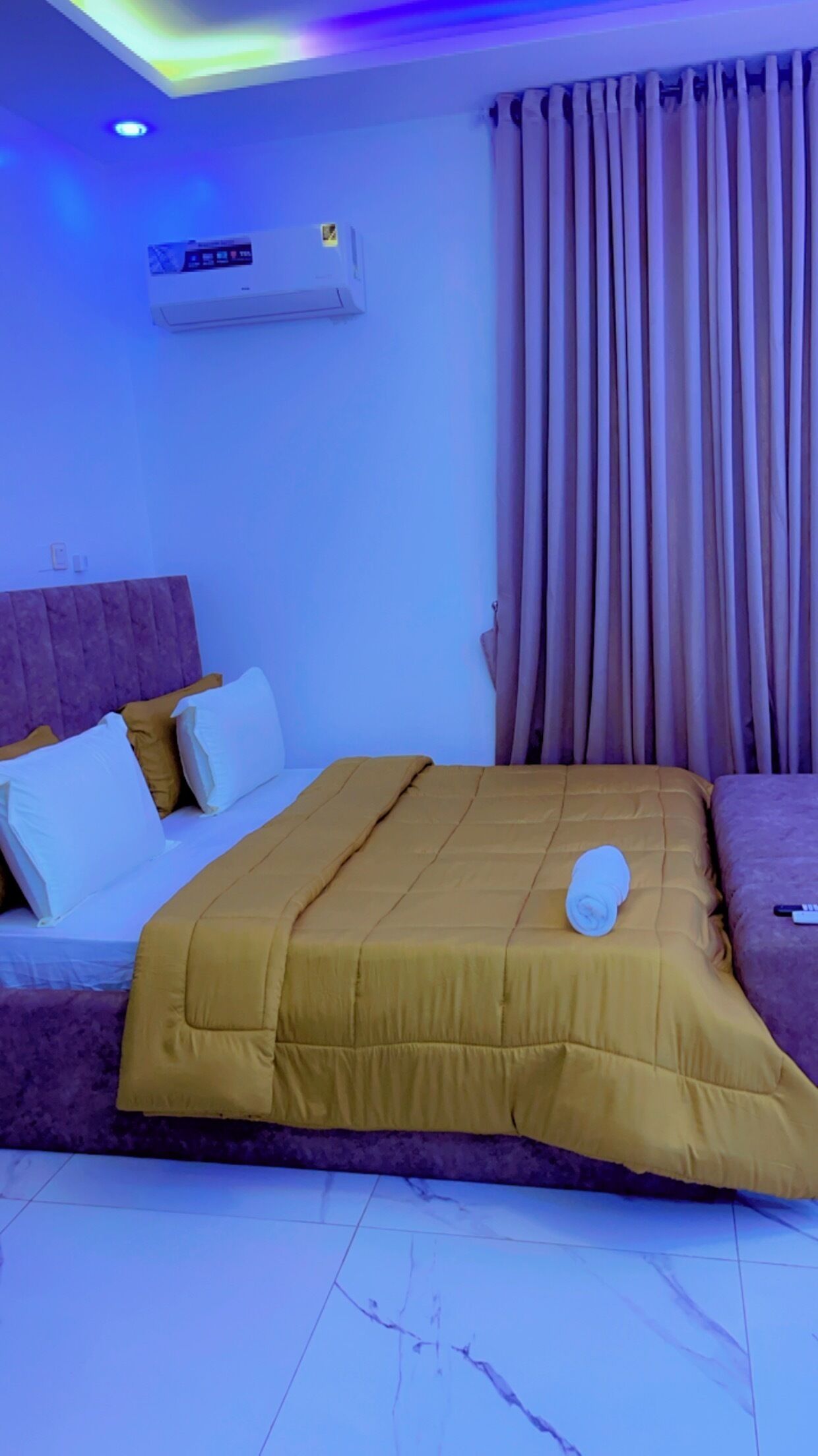 A hotel room with a bed, a headboard, a window with curtains, and an air conditioning unit.