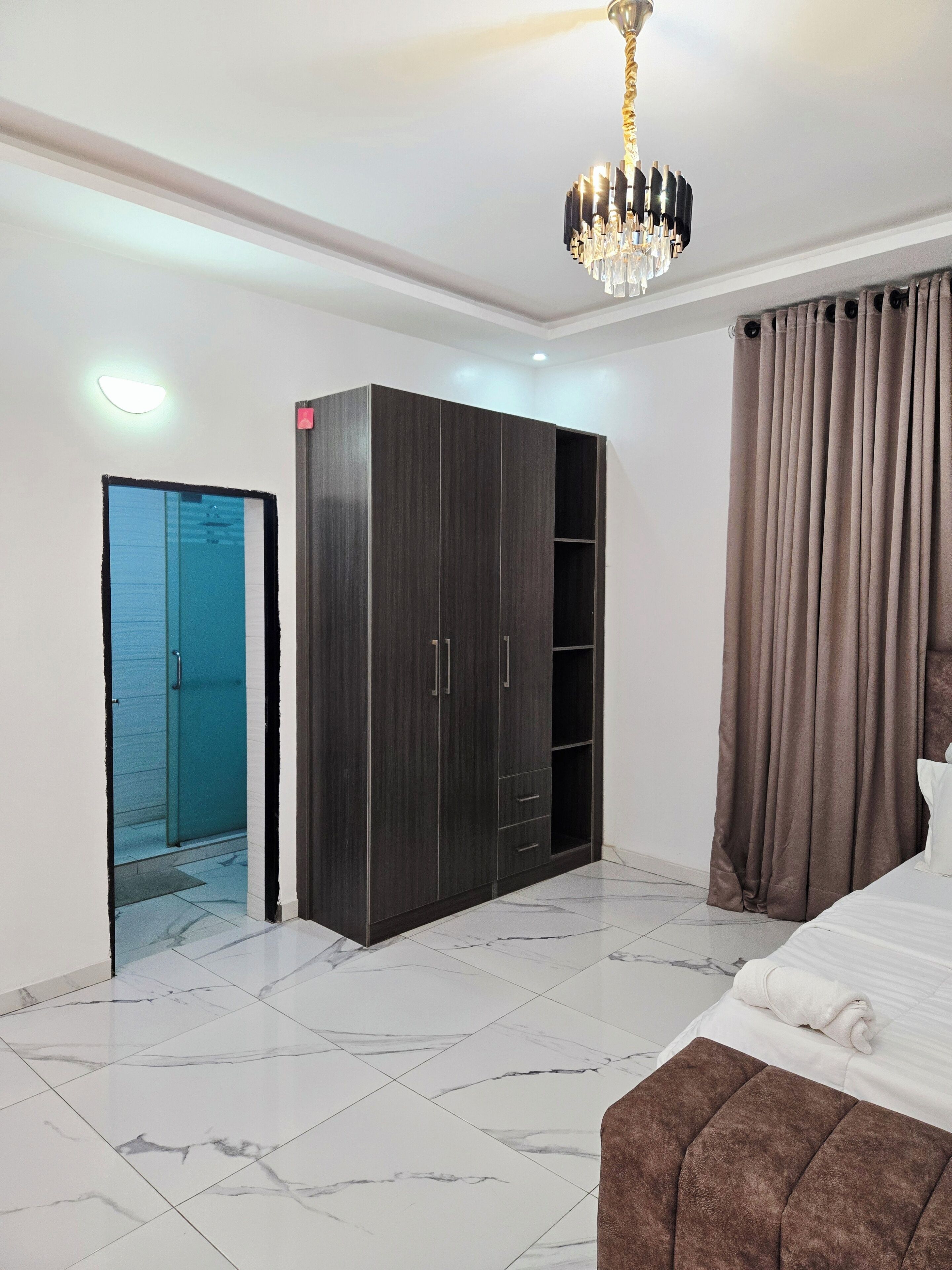 Quarto com cama, guarda-roupa, banheiro com porta de vidro e lustre.