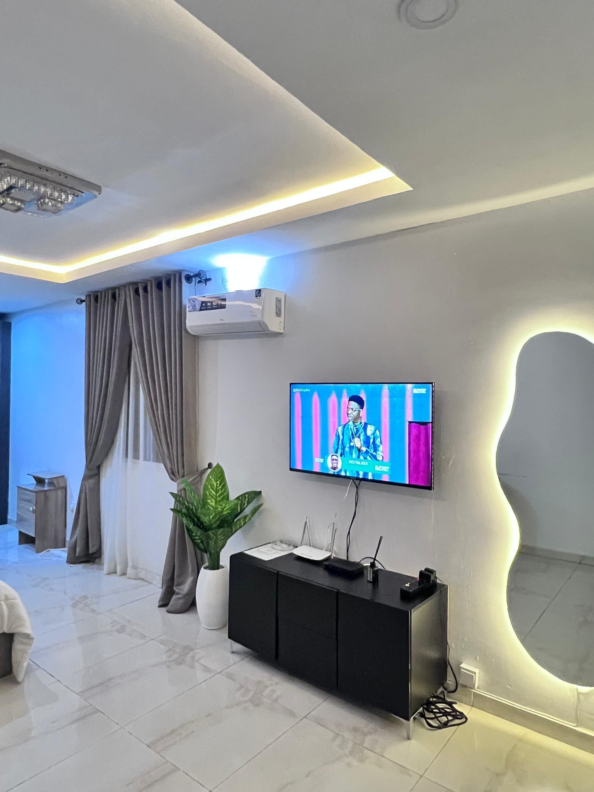 A modern living room with a flat-screen TV mounted on the wall, a black entertainment unit, a potted plant, and a ceiling light fixture.