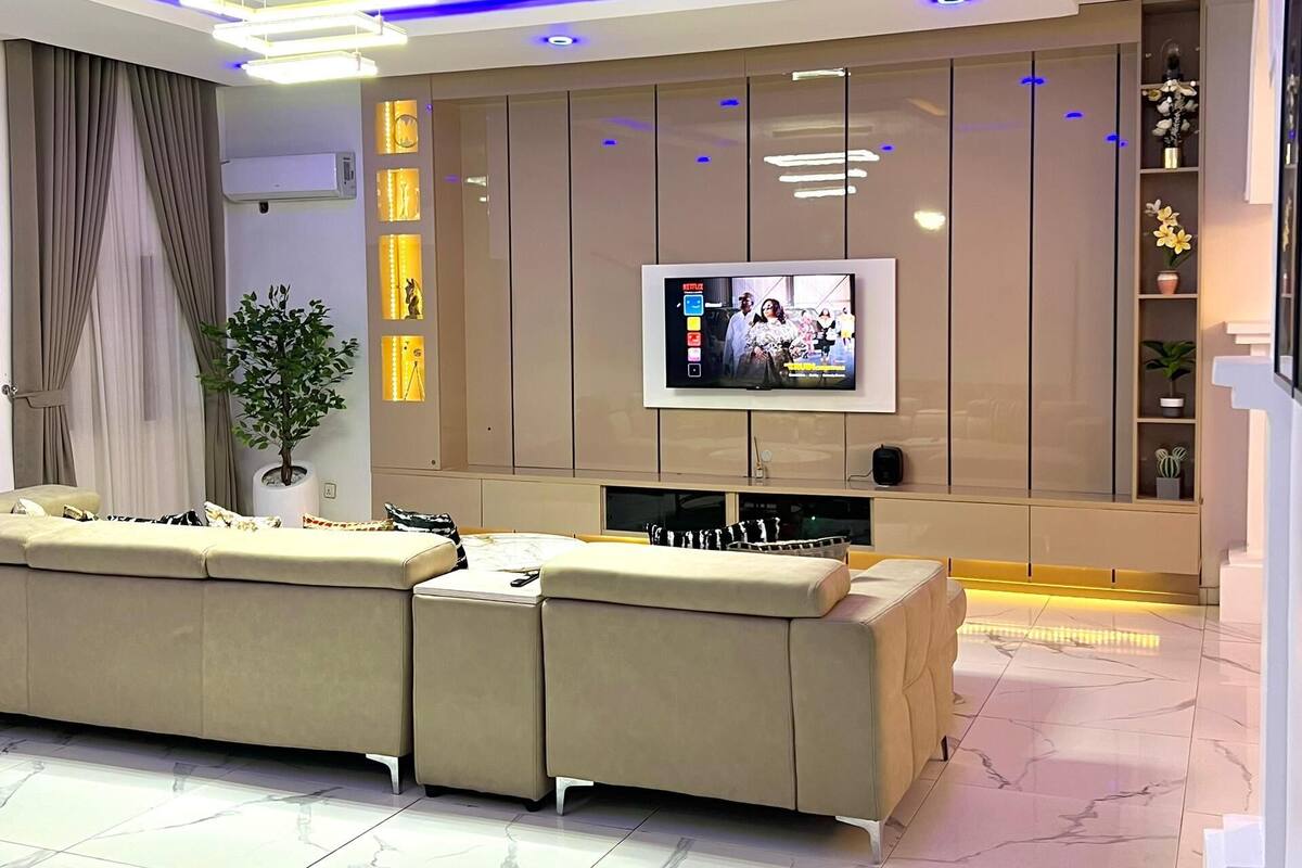 A modern living room with a large sectional sofa, a flat-screen TV mounted on a wall, and a potted plant.