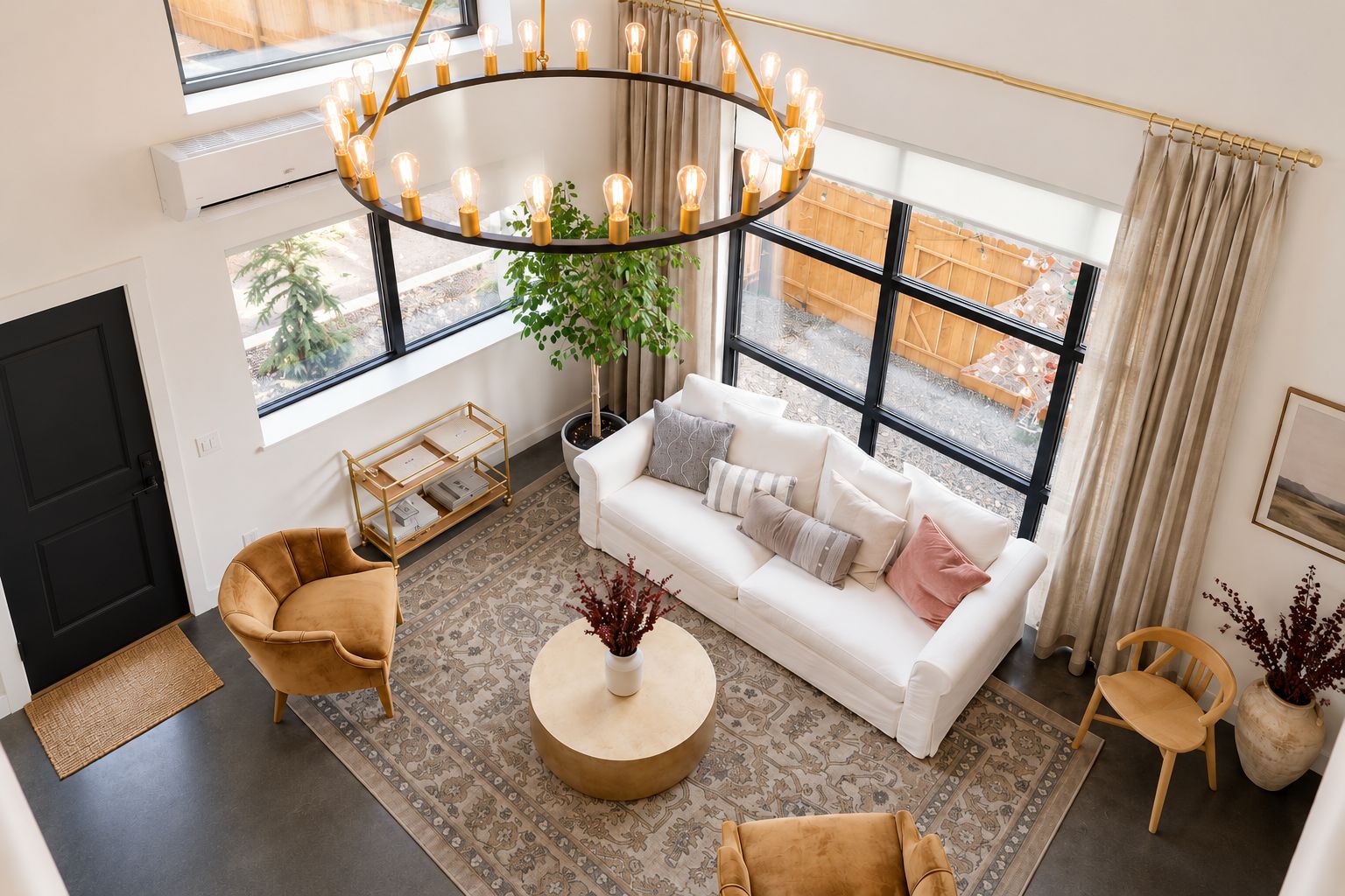 A modern living room with a white sofa, round coffee table, and two armchairs. A large window provides natural light, and a unique circular chandelier hangs above.