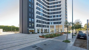 A modern building with a curved facade, a paved plaza with benches, and a street with parked cars. - Apartamento Malaga Towers - DolmenGroup (Málaga)