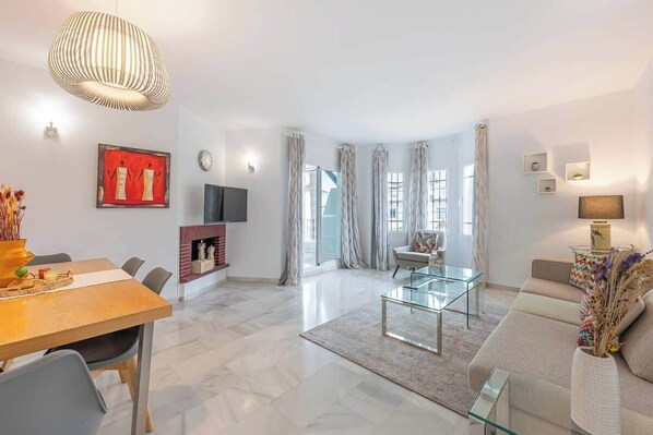 Un salon moderne avec une table basse en verre, un canapé, une table à manger avec des chaises, une cheminée et une grande fenêtre avec des rideaux.