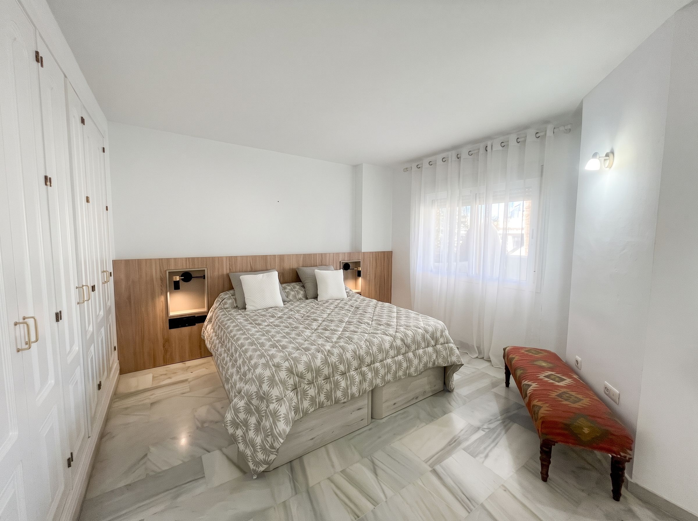 Une chambre à coucher avec un lit, des tables de chevet, un banc et une fenêtre avec des rideaux.