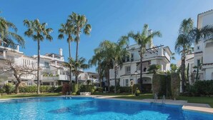 Una piscina rodeada de edificios blancos con balcones, palmeras y un cielo azul despejado.