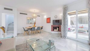 Una sala de estar moderna con una mesa de centro de vidrio, un comedor con sillas, una chimenea y un gran ventanal con cortinas.