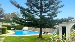Um quintal com piscina, uma grande árvore perene, um pequeno prédio branco e uma área cercada.