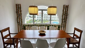 Uma área de refeições com uma mesa de madeira, cadeiras brancas e vista para um edifício através de uma janela.