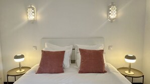 Cama com lençóis brancos e duas almofadas castanhas, com duas mesinhas de cabeceira com abajures e duas luminárias fixadas na parede.