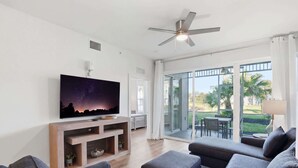 A modern living room with a flat-screen TV, grey sofas, a ceiling fan, and a sliding glass door leading to an outdoor patio. - #215 - First Floor - Sleeps 4- Landscape View (Naples)