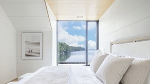 A bedroom with a large window, a bed with white bedding, and a framed picture on the wall. - Lake Joseph Beach house - Bringing the Caribbean to Muskoka (MacTier)