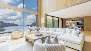 A modern living room with a large sofa, a coffee table, and a view of a lake. - Lake Joseph Beach house - Bringing the Caribbean to Muskoka (MacTier)