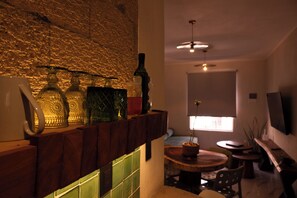 A room with a stone wall, a wooden shelf with glass bottles, a round table, and a TV mounted on the wall. - 15min from downtown,20min airport,5min walking cenote corazon & ZAMNA Festival. (Aldea Tulum)