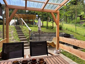 Ein überdachter Außenbereich mit Holztisch, zwei Stühlen und Blick auf einen Garten sowie eine Holzkonstruktion im Hintergrund.
