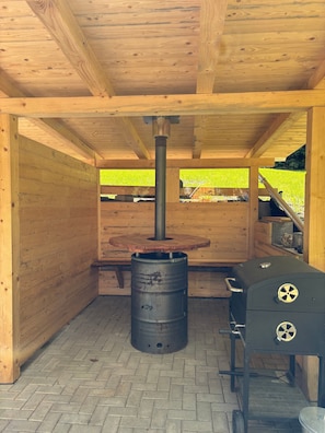 Ein überdachter Außenbereich mit Kaminofen, Grill und einer Sitzbank.